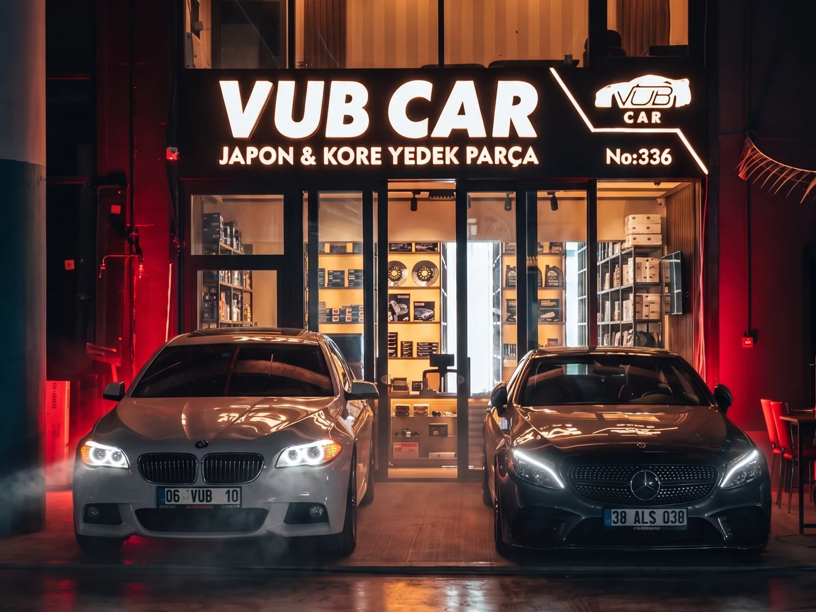 VUB CAR mağazasının dış cephesi: aydınlatmalı Japon ve Kore yedek parça tabelası, vitrinde kutulu parça rafları, önünde park etmiş iki araç